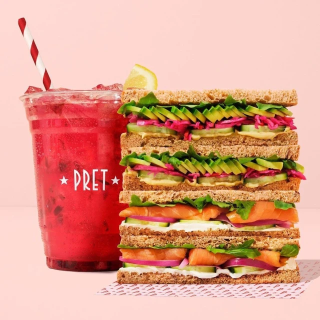 Tall sandwich stacked with smoked salmon, avocado, cucumber, and pink pickled onions, served with a vibrant berry drink with a straw.
