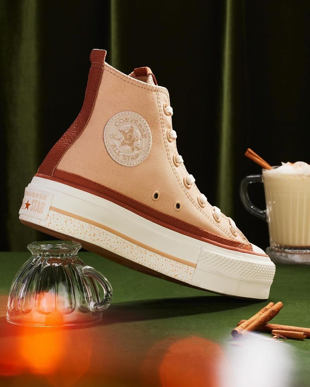 High-top Converse sneaker with a tan and brown color scheme, displayed on a green surface with a glass and a drink.