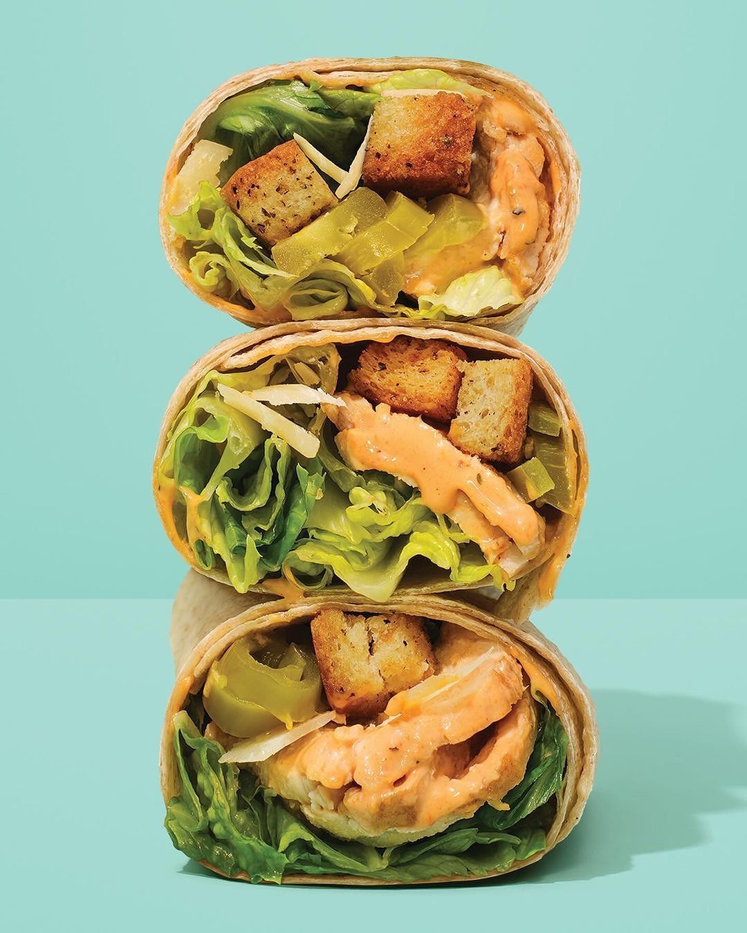 Three stacked wrap sandwiches filled with lettuce, shredded cheese, croutons, and a creamy sauce against a light blue background.
