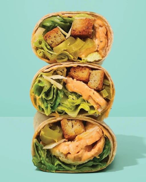 Three stacked wrap sandwiches filled with lettuce, shredded cheese, croutons, and a creamy sauce against a light blue background.