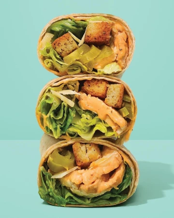 Three stacked wrap sandwiches filled with lettuce, shredded cheese, croutons, and a creamy sauce against a light blue background.