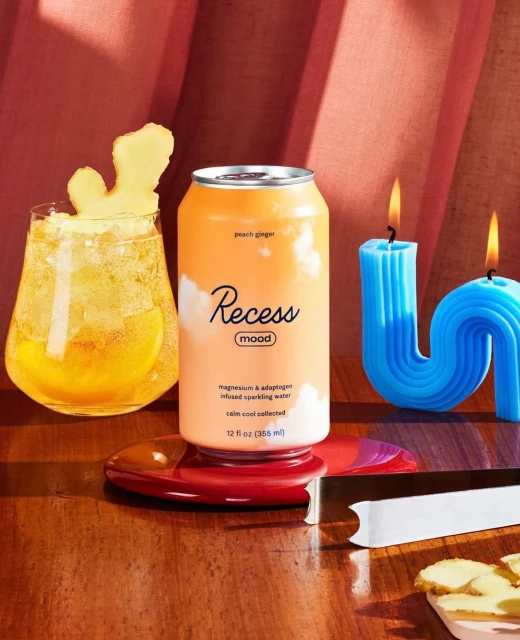 Recess Mood sparkling water can and glass of ginger ale drink with ginger slice, displayed with blue candles on a wood table.