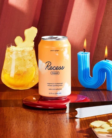 Recess Mood sparkling water can and glass of ginger ale drink with ginger slice, displayed with blue candles on a wood table.
