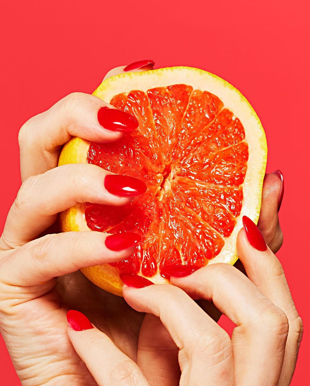 Close-up of a half grapefruit held in two hands with red nail polish against a vibrant red background. Food and beauty concept.