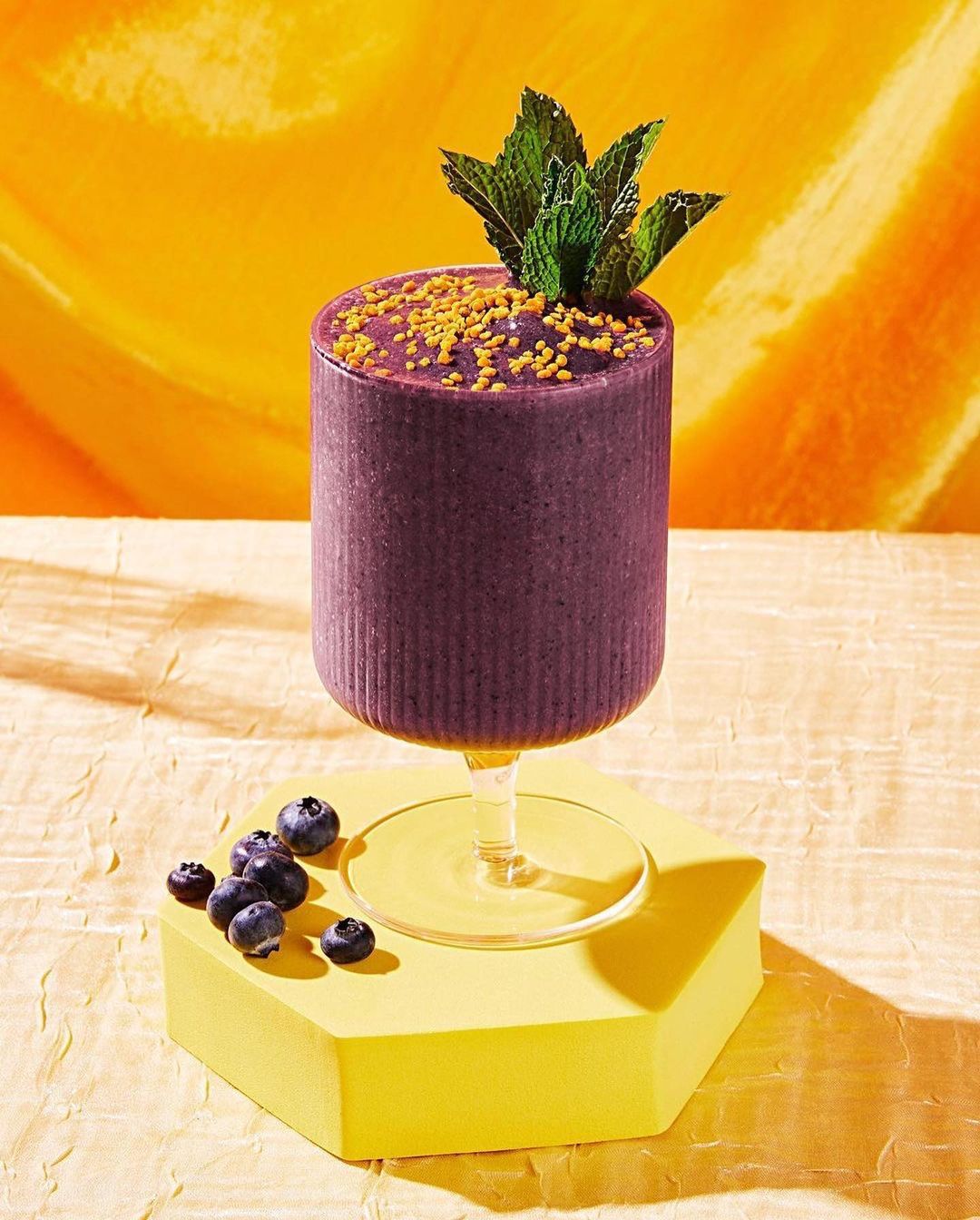 A vibrant purple smoothie in a stemmed glass, topped with mint and yellow granules, set against a textured yellow backdrop.