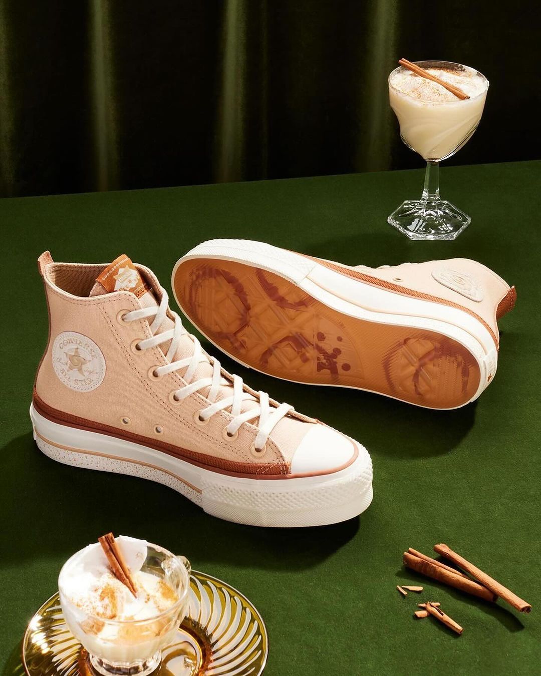 Pair of platform Converse sneakers are staged near two dessert cocktails garnished with cinnamon sticks, against a green background.