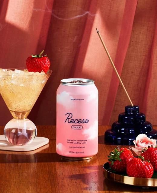 Recess mood sparkling water can alongside cocktail glass with strawberry garnish, incense holder, and strawberries on a wooden table.