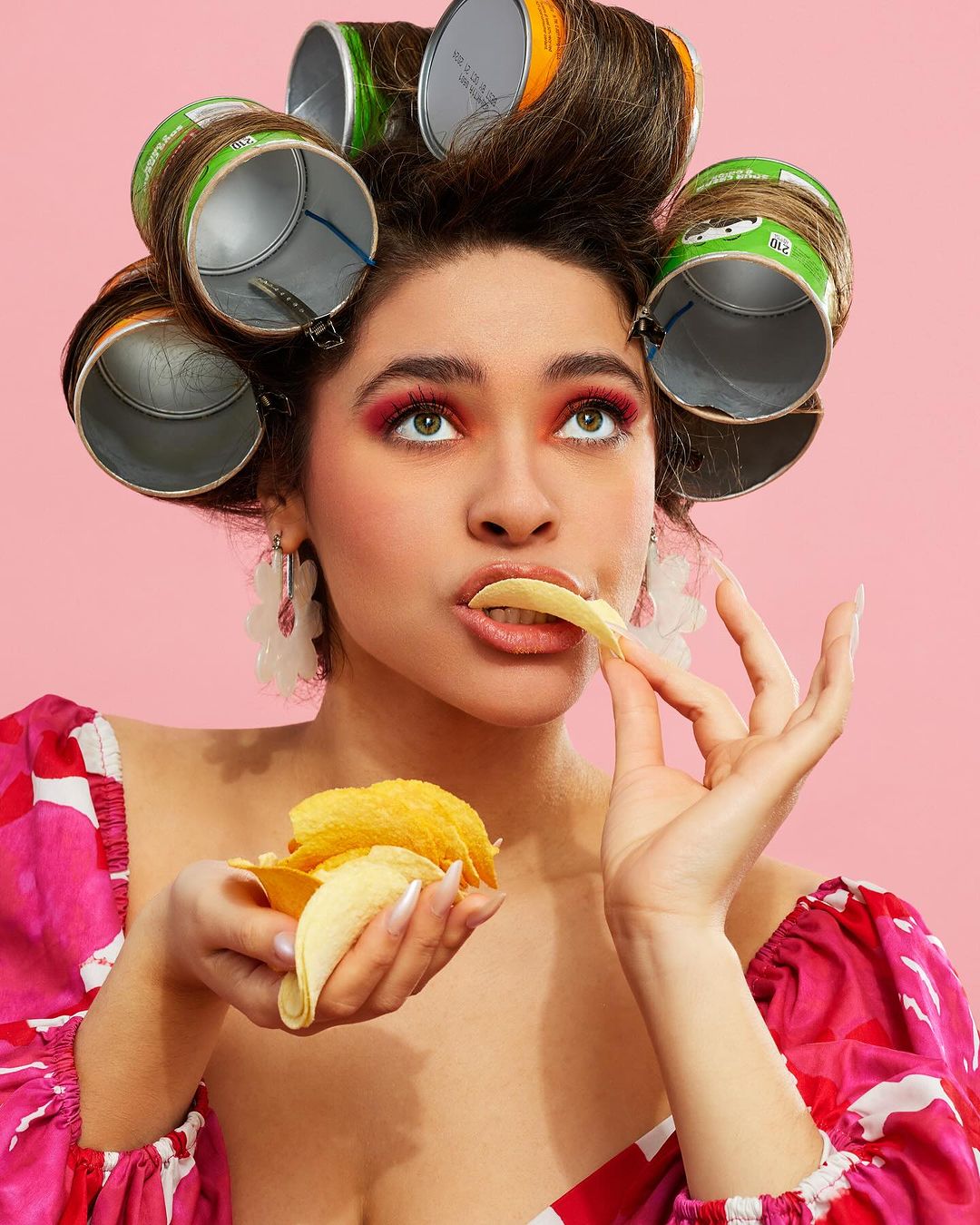 A young woman with Pringles cans as hair rollers eats a potato chip in front of a pink backdrop, in a beauty and food-themed editorial shot.