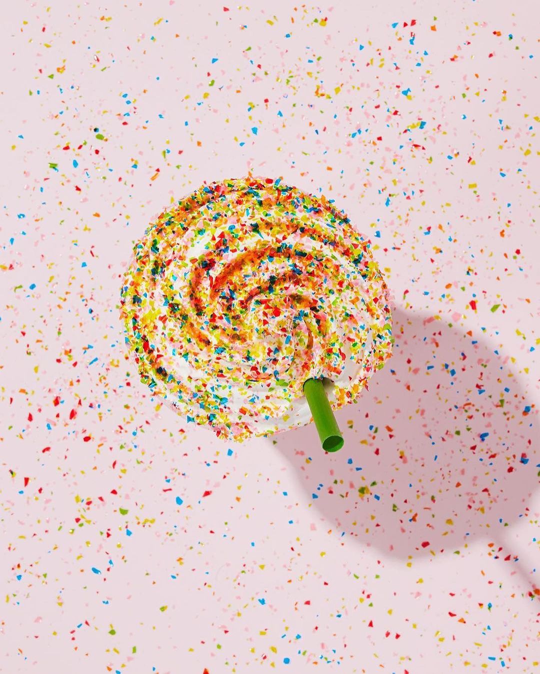Overhead shot of a white frosted dessert or drink with colorful sprinkles and a green straw against a light pink background.