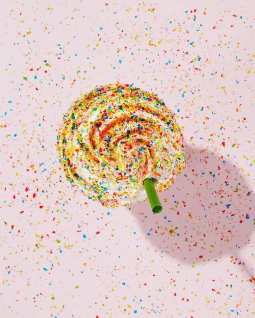 Overhead shot of a white frosted dessert or drink with colorful sprinkles and a green straw against a light pink background.