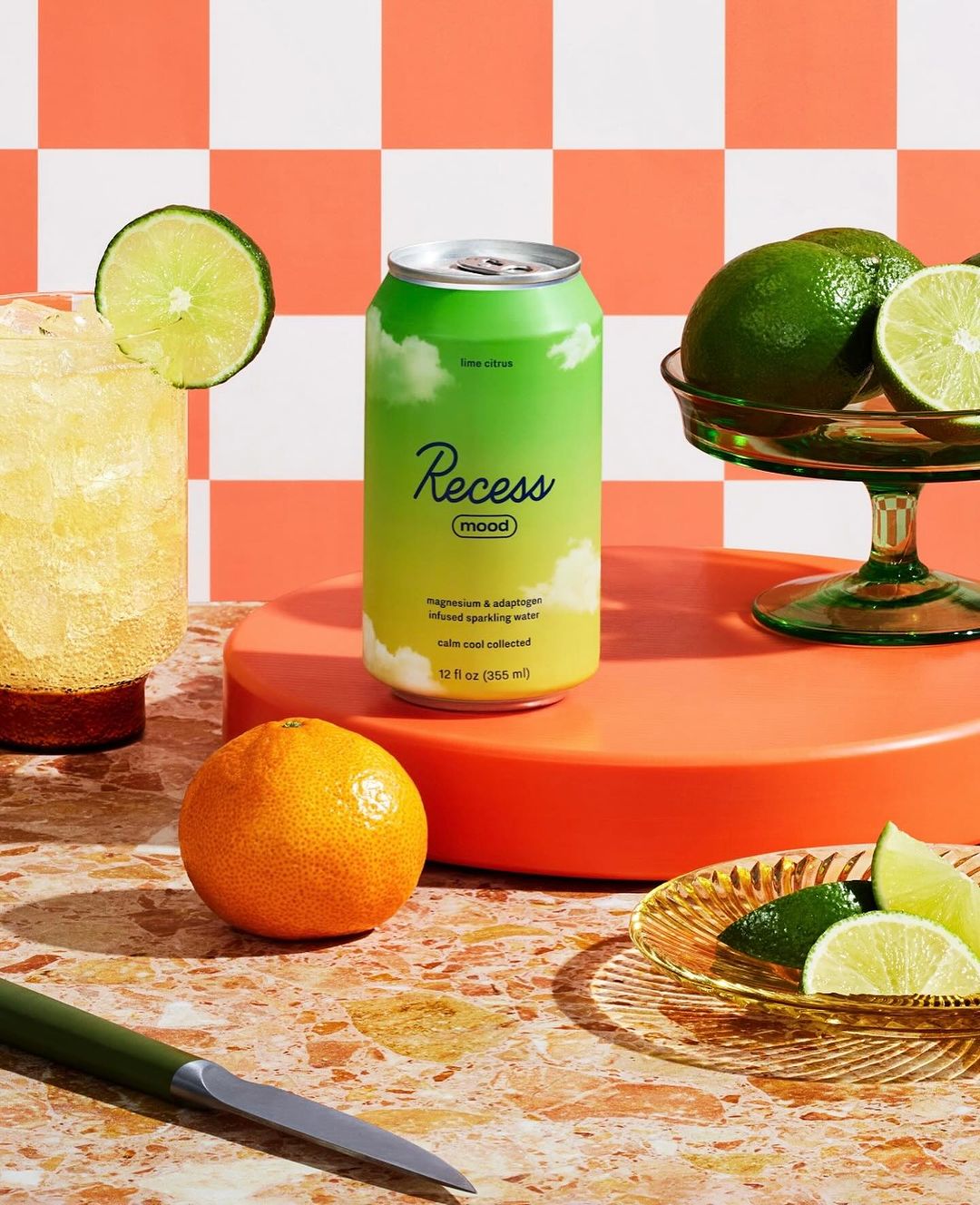 Still life of a can of Recess sparkling water surrounded by limes, mandarins, and a cocktail against an orange and white checkered background.