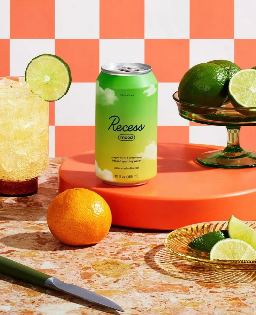 Still life of a can of Recess sparkling water surrounded by limes, mandarins, and a cocktail against an orange and white checkered background.