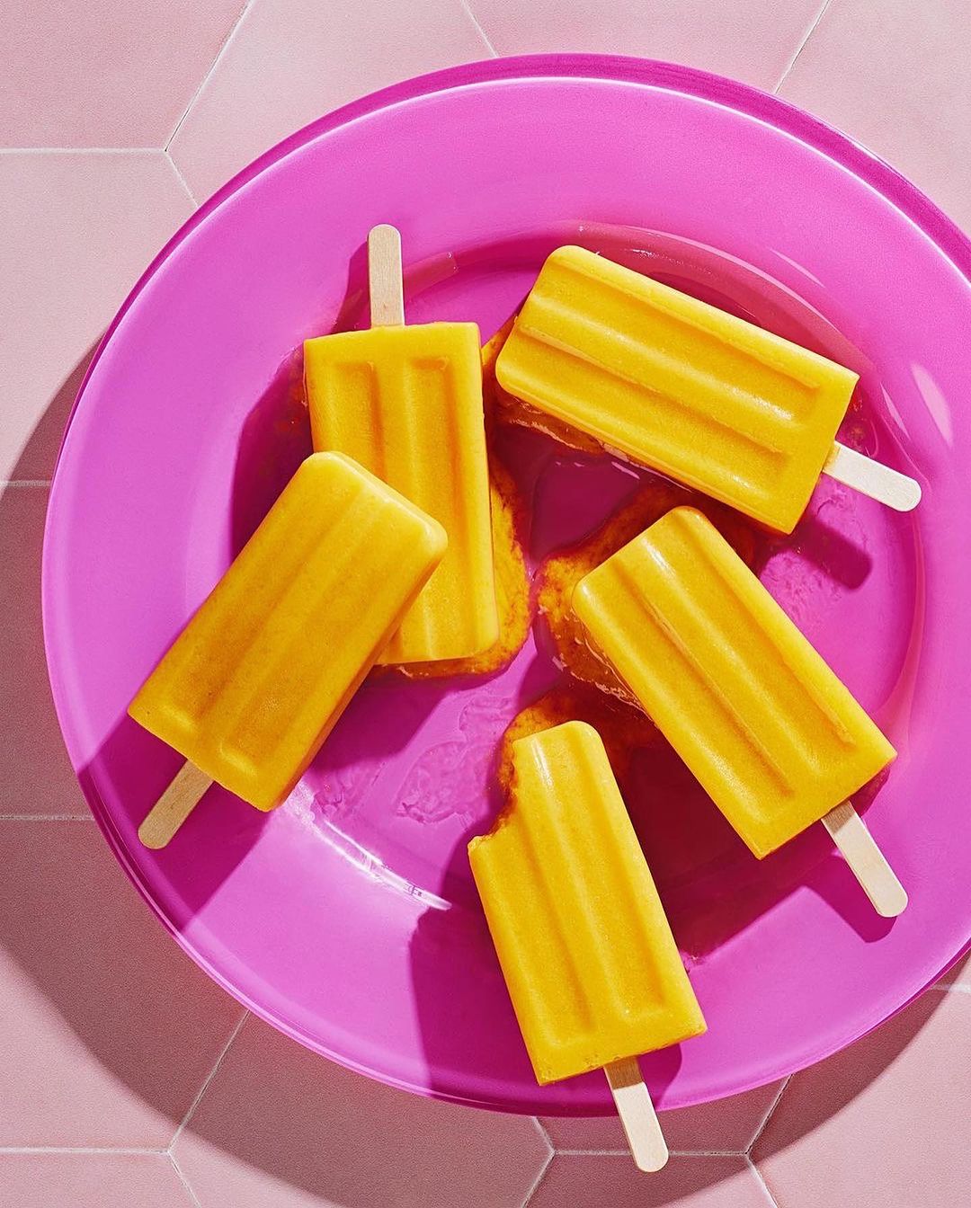 Five yellow popsicles on a pink plate, arranged on a pink hexagonal tile surface, with melted drips adding to the vibrant, summery scene.