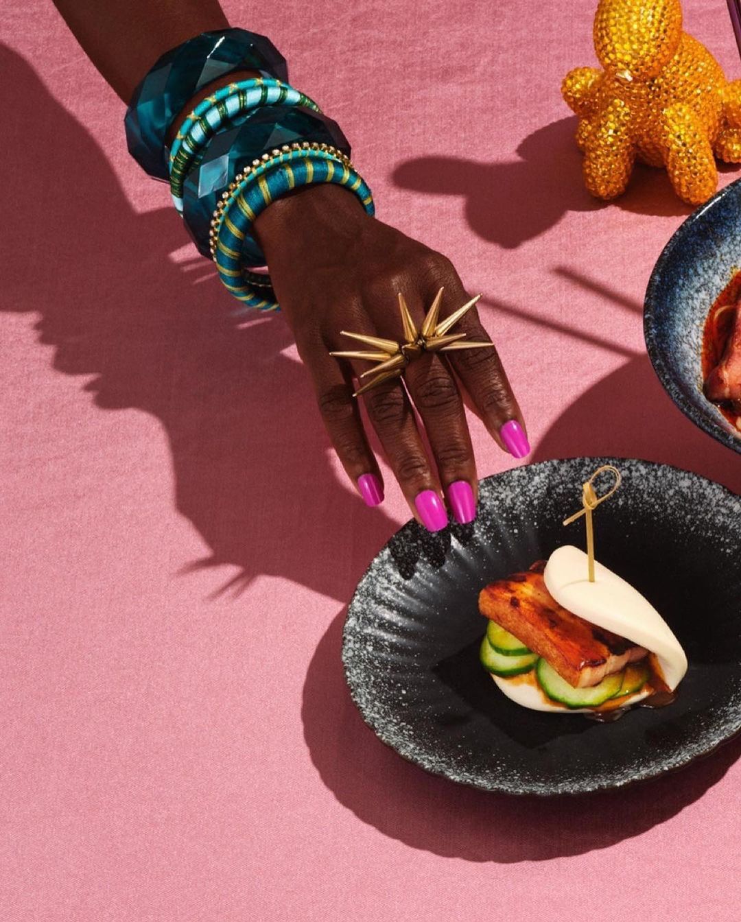 Close-up of a hand with an exotic ring, bracelets, and bright pink nail polish, near a plate with a pork bun.