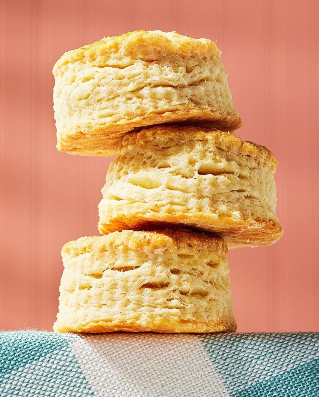 Close-up of a stack of three golden-brown biscuits on a blue and white checkered cloth against a solid light-red background.