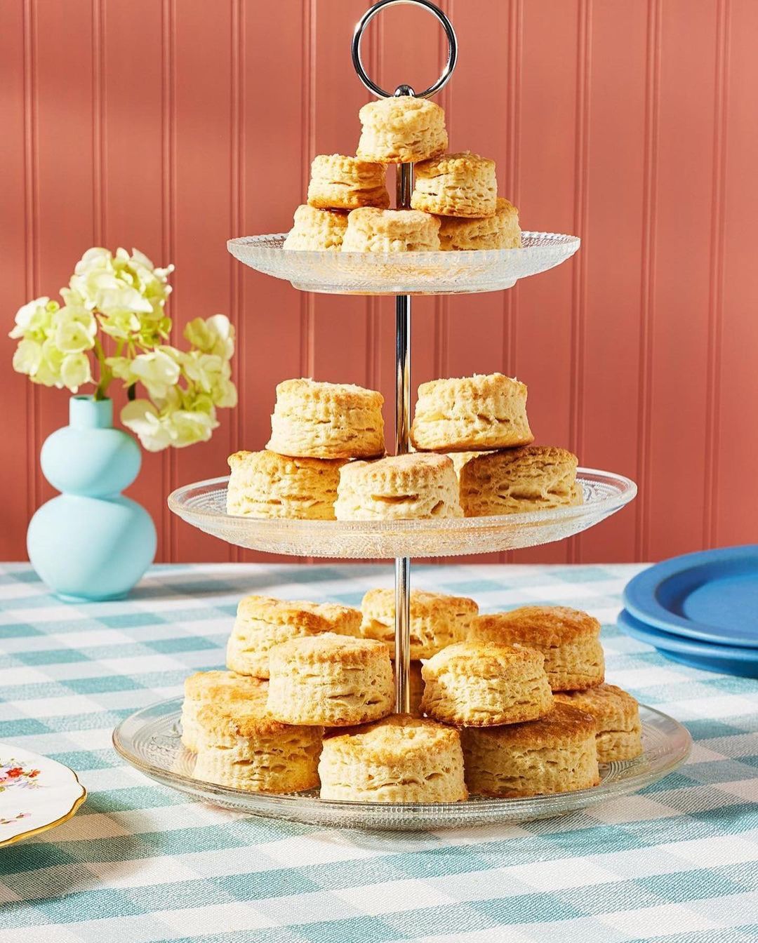 Three-tiered dessert stand filled with scones on a gingham tablecloth next to a vase of flowers, with a peach wall in the background.