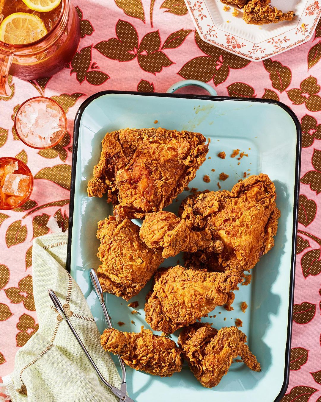 Overhead shot of golden fried chicken on a blue platter, accompanied by iced tea and tongs, set against a floral tablecloth.