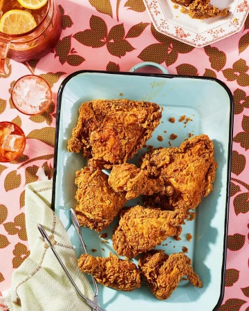 Overhead shot of golden fried chicken on a blue platter, accompanied by iced tea and tongs, set against a floral tablecloth.