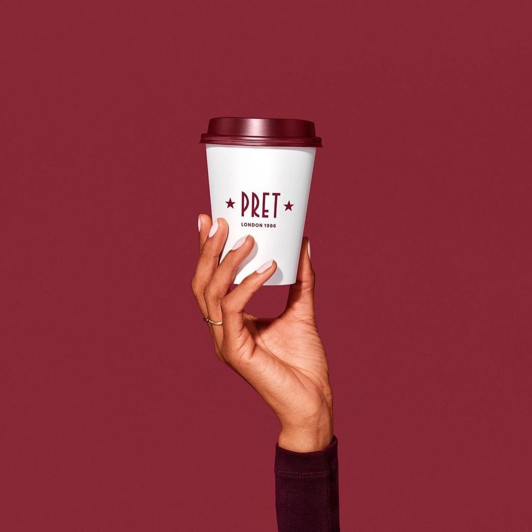 A woman's hand holds a disposable coffee cup with the Pret a Manger logo on a maroon background, showcasing a simple and clean aesthetic.