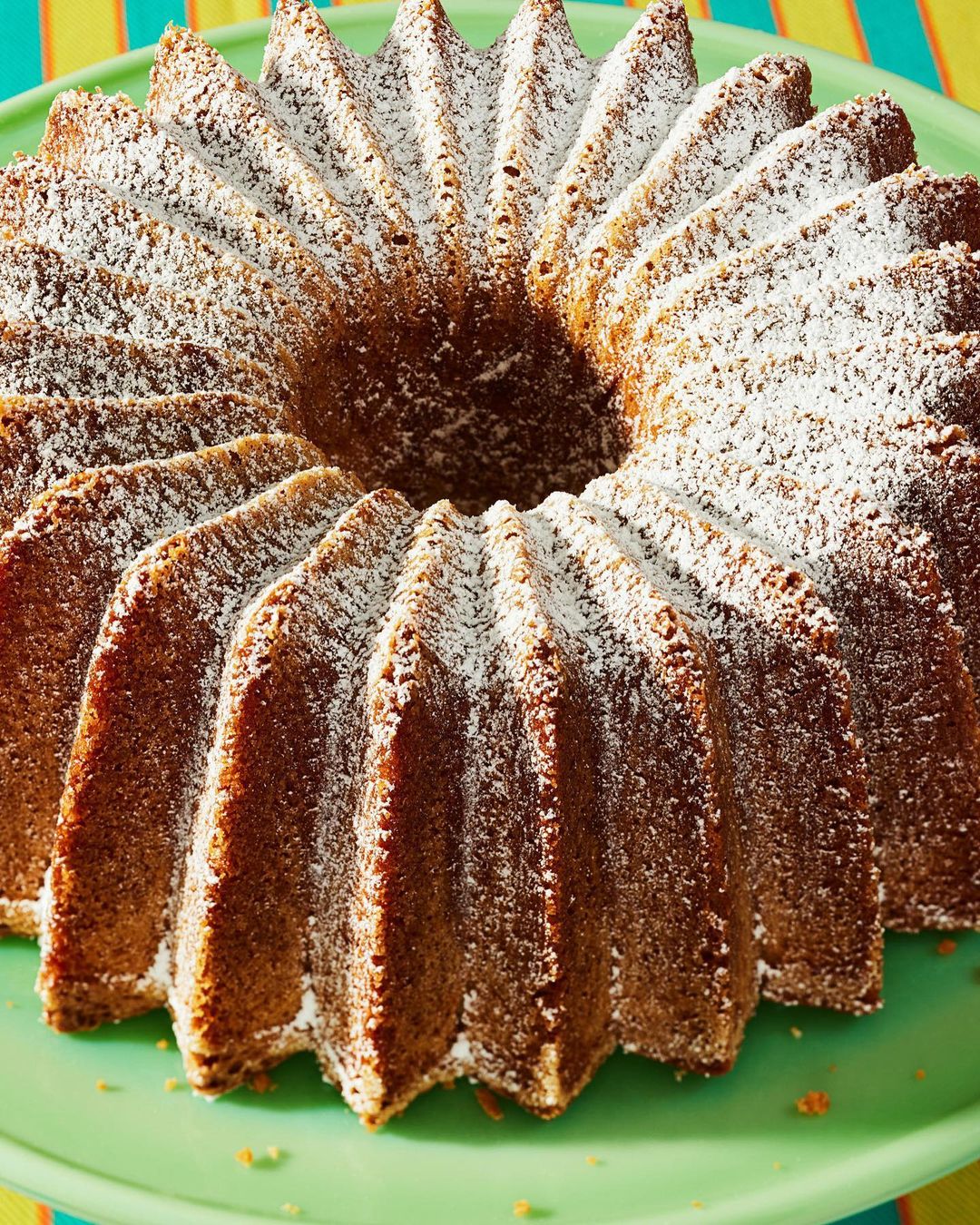 Bundt cake covered in powdered sugar on a green cake stand, with a colorful striped background out of focus.