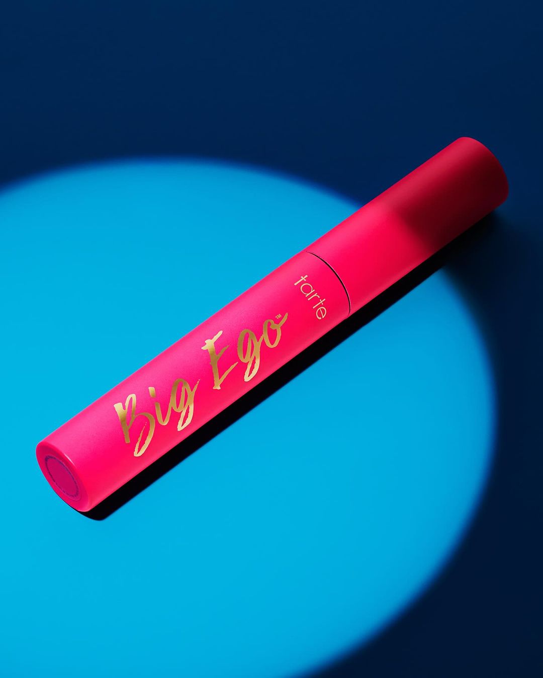 Tarte Big Ego mascara tube in hot pink with gold lettering on a bright blue background with lighting effects.