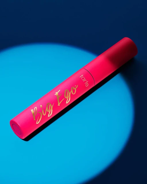 Tarte Big Ego mascara tube in hot pink with gold lettering on a bright blue background with lighting effects.