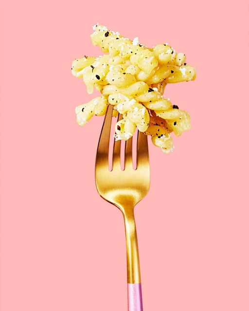 Close-up of spiral pasta with cheese and black pepper, plated on a gold-colored fork against a light pink background.