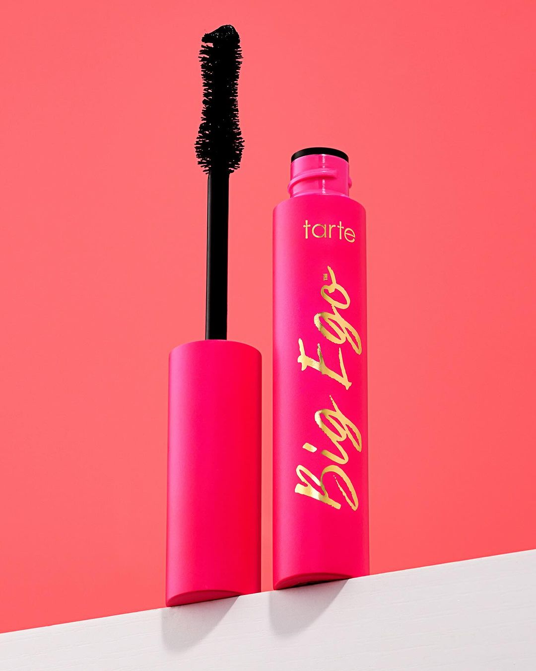 Tarte Big Ego mascara in a pink tube with a black wand, displayed on a white surface against a coral pink backdrop.