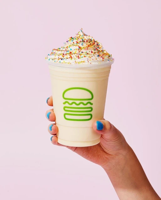 Hand holding vanilla milkshake topped with sprinkles in a clear plastic cup against a pink background, focus on the creamy dessert.