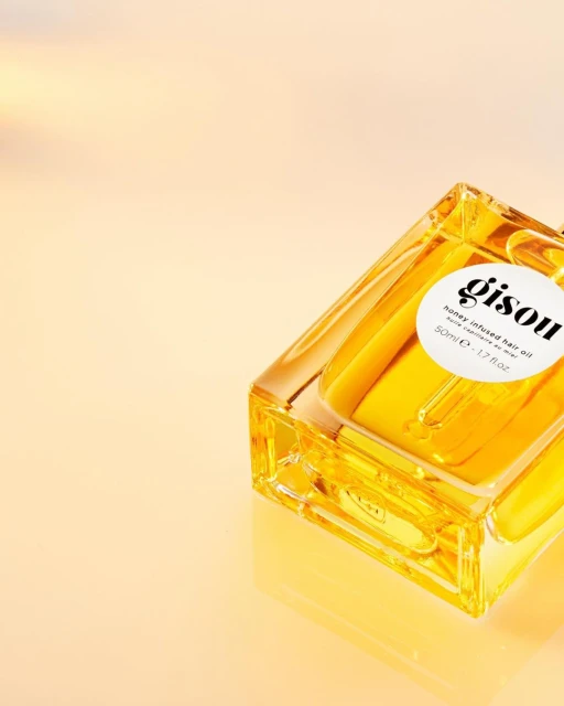 Gisou honey-infused hair oil in a clear glass bottle with a label, showcasing its golden color against a gradient orange background.
