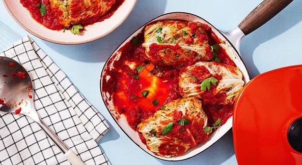 Overhead view of cabbage rolls in tomato sauce in a white pan with an orange lid, alongside a check towel and spoon.