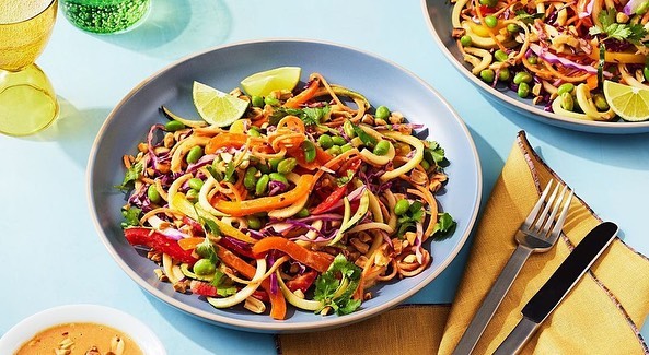Spiralized vegetable noodle salad with red cabbage, edamame, and lime wedge served in a blue bowl, alongside silverware and a small bowl of sauce.