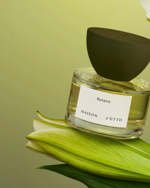 Close-up of a Maison d'Etto Rotano perfume bottle with a dark green cap sitting on lily petals against a light green background.