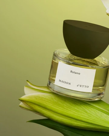 Close-up of a Maison d'Etto Rotano perfume bottle with a dark green cap sitting on lily petals against a light green background.