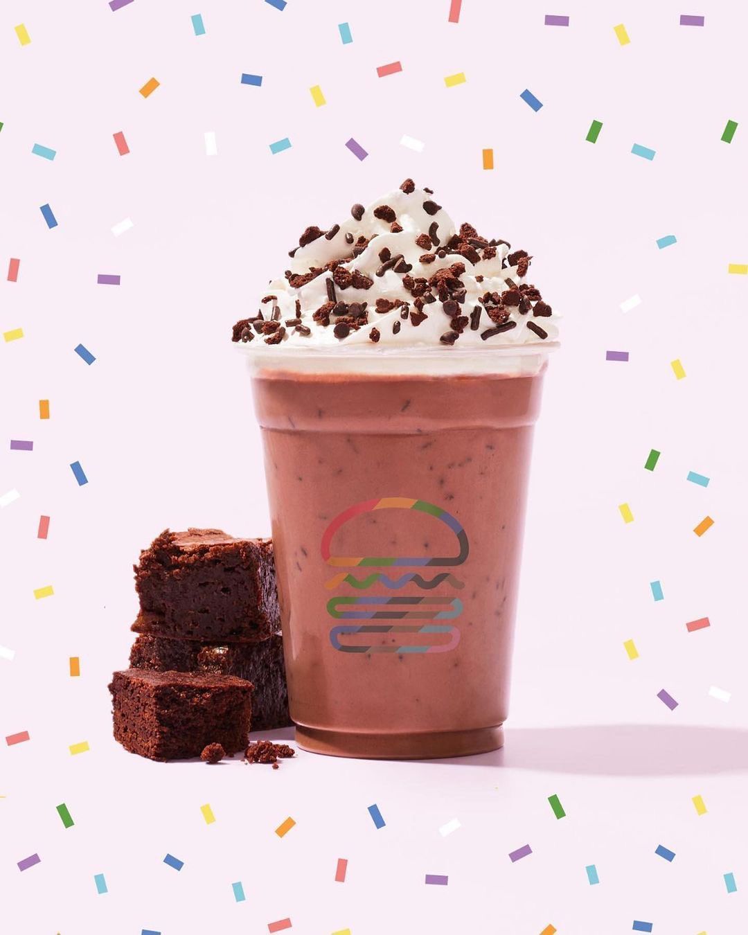 A chocolate milkshake in a clear plastic cup, topped with whipped cream and chocolate sprinkles, next to a stack of chocolate brownies.