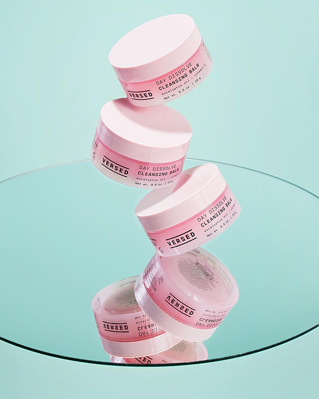 Stack of Versed Day Dissolve Cleansing Balm product containers against a light teal background, reflected in a mirror surface.