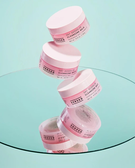 Stack of Versed Day Dissolve Cleansing Balm product containers against a light teal background, reflected in a mirror surface.