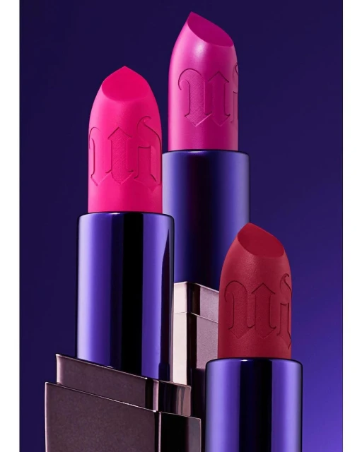 Three tubes of lipstick in bright hues of pink and red stand against a graduated blue background for a beauty product shot.