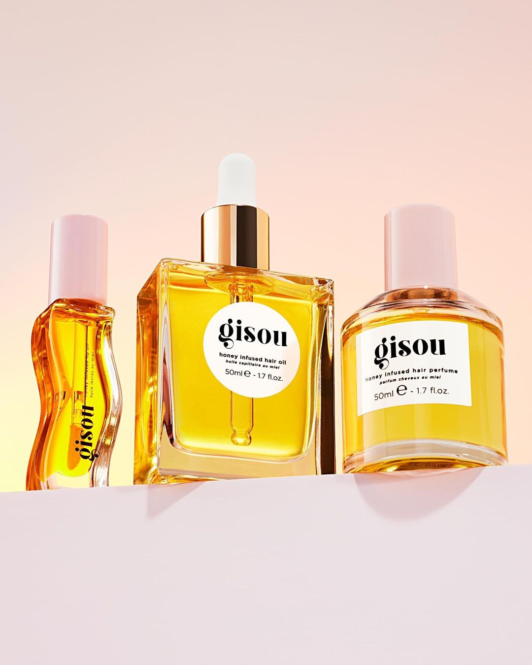 Three Gisou hair care products, honey-infused hair oil and perfume, line a pink shelf against a soft orange and pink gradient background.