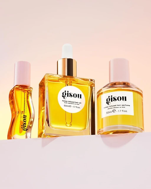 Three Gisou hair care products, honey-infused hair oil and perfume, line a pink shelf against a soft orange and pink gradient background.