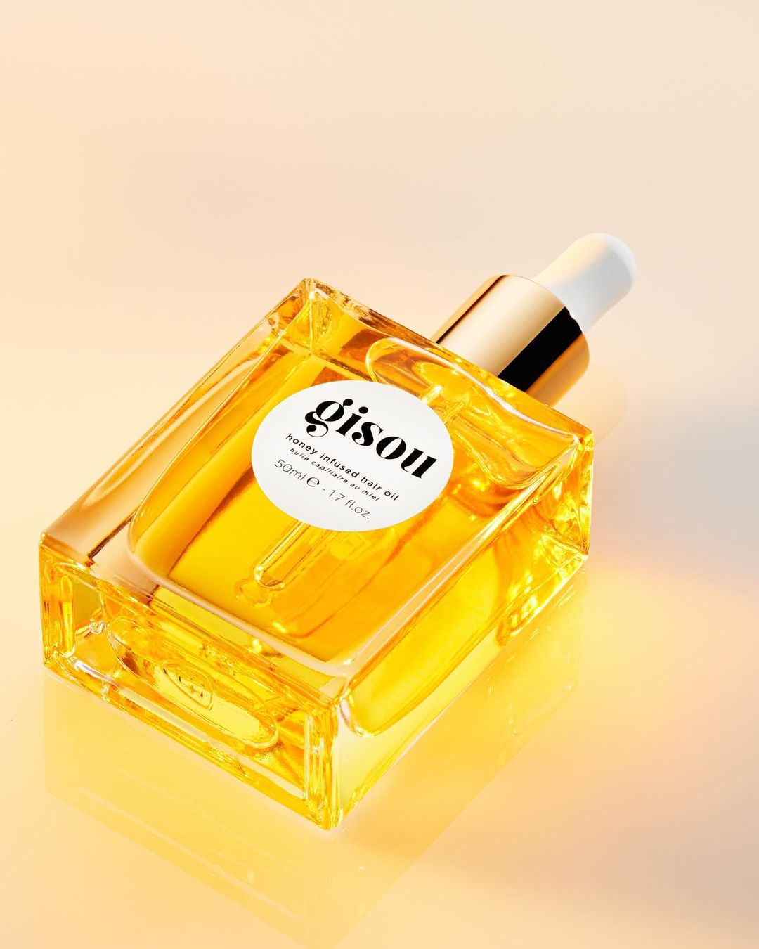 Studio shot of Gisou honey infused hair oil in a clear square bottle with a white labeled circle logo on a peach gradient background.