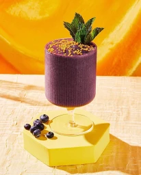 A purple smoothie in a ribbed glass topped with mint leaves and seeds, resting on a yellow hexagonal pedestal with scattered blueberries.