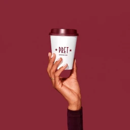 Hand holding a white Pret a Manger takeaway coffee cup with burgundy lid against a solid burgundy background