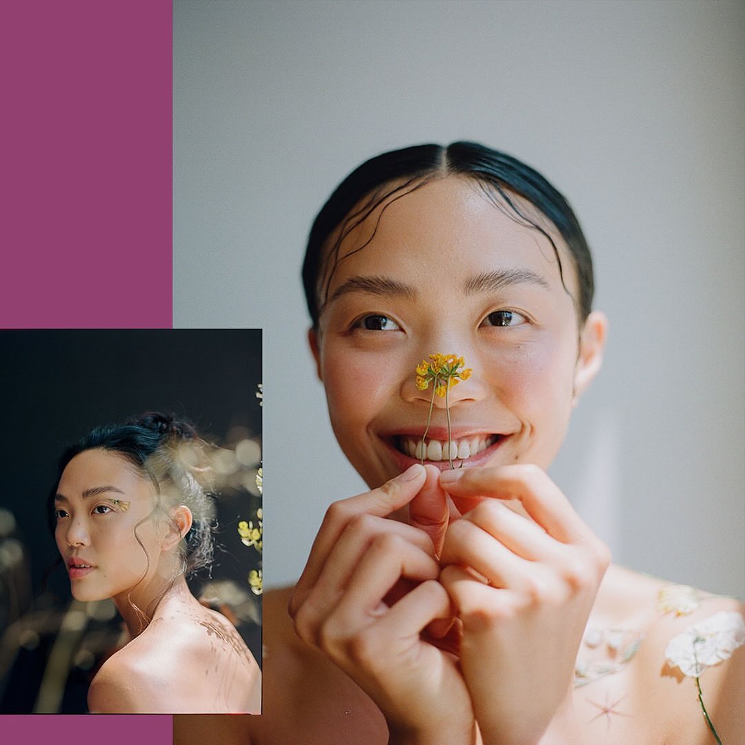 Close-up portrait of a smiling Asian woman with a flower on her nose, artistic makeup, and dewy skin, against a light gray background.
