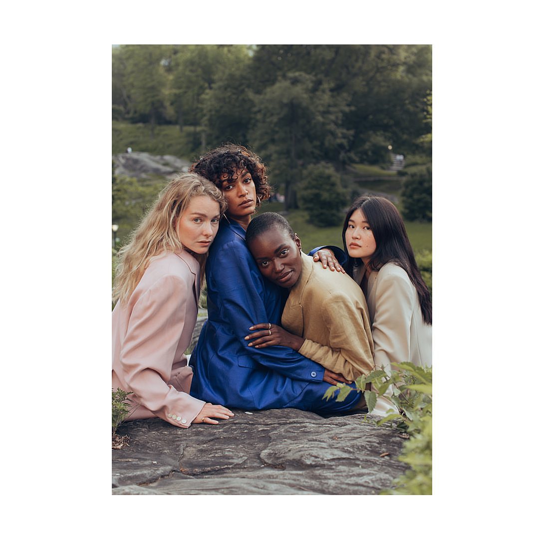 A group of four multiethnic women in stylish blazers pose closely together against a backdrop of lush green trees in an outdoor setting.