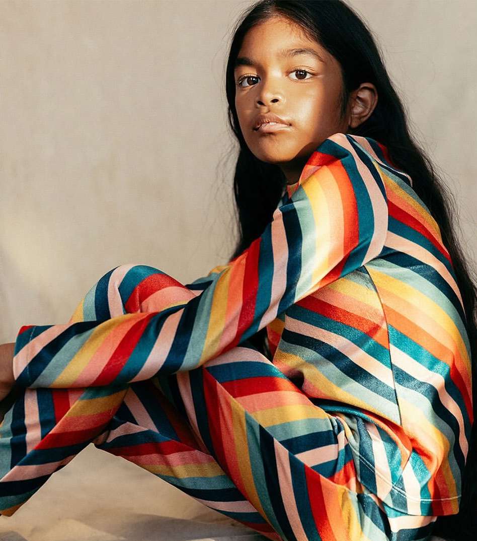 A young child with long hair poses indoors, wearing a colorful striped blazer and pants suit against a neutral background in soft light.