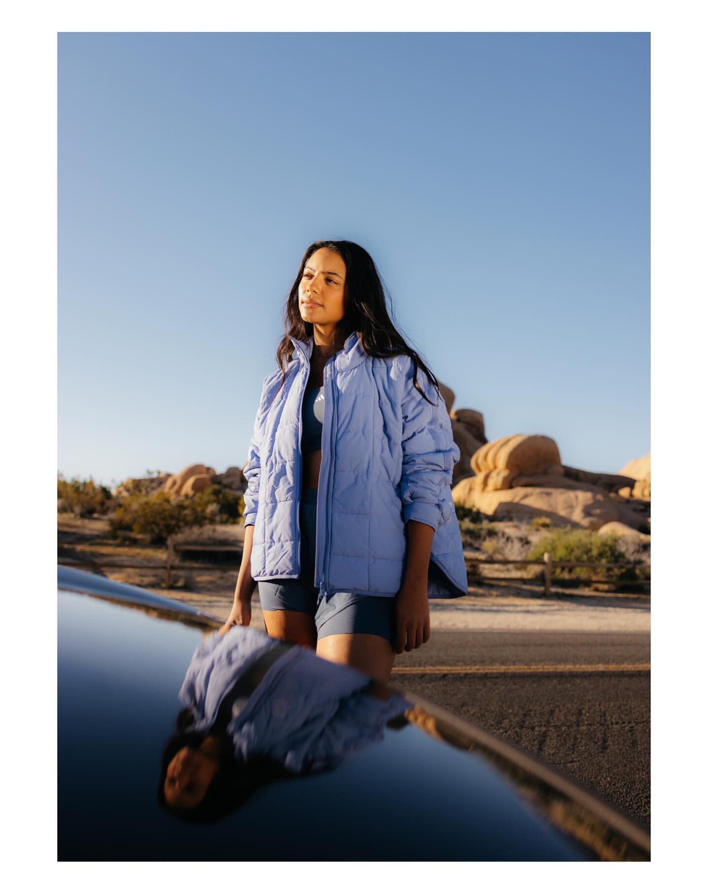 A woman wearing a light blue jacket and shorts poses beside a car, with desert rocks and a clear sky in the background.