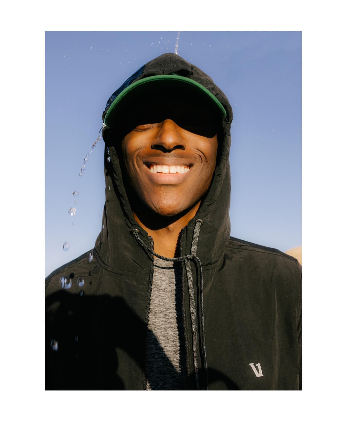 Smiling African-American man in a hooded jacket and baseball cap, with water droplets falling, set against a clear blue sky backdrop.