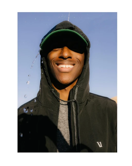 Smiling African-American man in a hooded jacket and baseball cap, with water droplets falling, set against a clear blue sky backdrop.