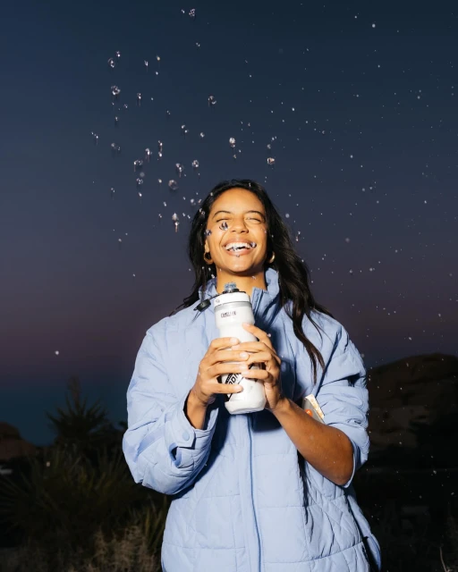 Woman in light blue jacket smiles as water is splashed on her. She holds a white water bottle in an outdoor dusk environment.